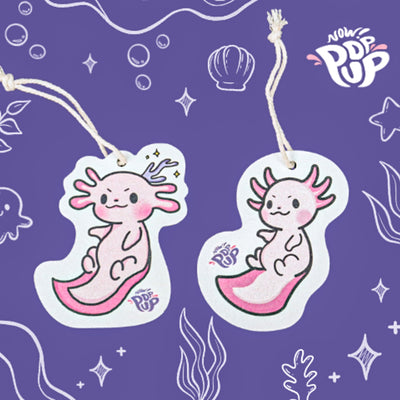 Top view of an eco-friendly cellulose pop-up kitchen sponge with a hanging cord, featuring a pattern of two adorable axolotls and engraved with the brand name "now pop up"—compact when dry and expanding when wet for cheerful cleaning