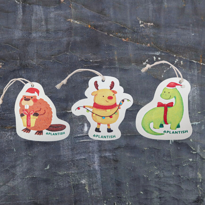 Top view of three eco-friendly pop-up sponges featuring festive holiday animal designs. This charming set includes: 1) Holiday Moose, 2) Holiday Beaver, and 3) Holiday Dinosaur patterns. Displayed on a dark grey textured background, these circular kitchen sponges are crafted from biodegradable cellulose with attached strings, bringing cheerful holiday spirit to your cleaning routine while being kind to the environment