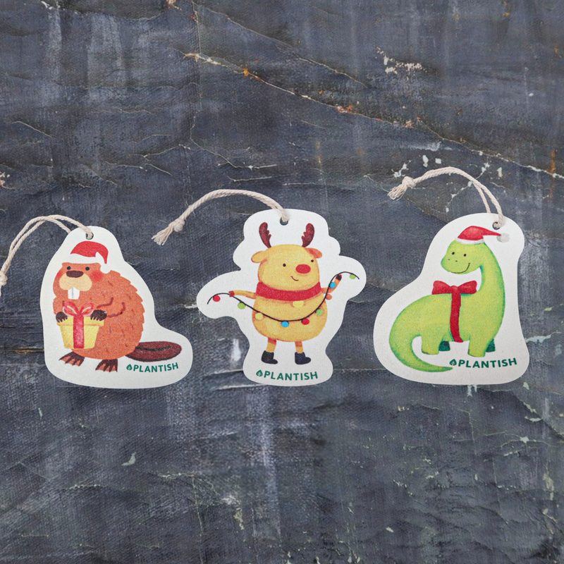 Top view of three eco-friendly pop-up sponges featuring festive holiday animal designs. This charming set includes: 1) Holiday Moose, 2) Holiday Beaver, and 3) Holiday Dinosaur patterns. Displayed on a dark grey textured background, these circular kitchen sponges are crafted from biodegradable cellulose with attached strings, bringing cheerful holiday spirit to your cleaning routine while being kind to the environment