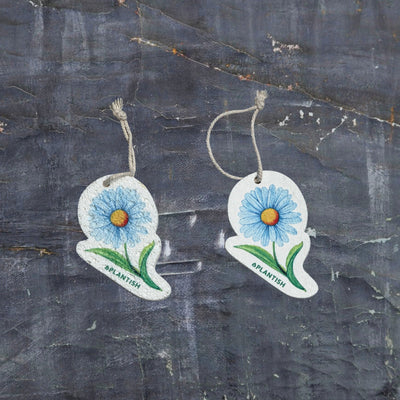 A top-view comparison showing the Blue Daisy pop-up sponge in dry versus wet states. On the right: the sponge appears flat and compact in its dry form. On the left: it has expanded into a thicker cleaning pad after hydration. Both clearly display the charming blue daisy pattern and feature an attached hanging string. Photographed against a dark grey textured background, this eco-friendly kitchen cleaner demonstrates remarkable size expansion when water is absorbed.