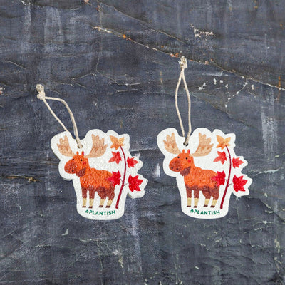 This top-view comparison shows the Autumn Moose pop-up sponge in dry (right: flat/compact) versus wet (left: expanded/thicker) states, clearly displaying its red moose and maple tree design with hanging cord against a dark grey textured background, demonstrating the eco-friendly cleaner's remarkable water-activated expansion.