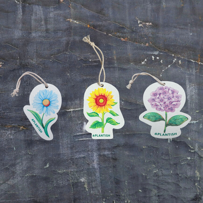 Top view of three eco-friendly pop-up sponges featuring beautiful floral designs. The set includes: 1) Sunflower, 2) Purple hydrangea, and 3) Blue daisy patterns. These circular kitchen sponges are displayed on a dark grey textured background. Made from biodegradable cellulose with attached strings, they combine practical cleaning with the beauty of nature in your everyday routine.