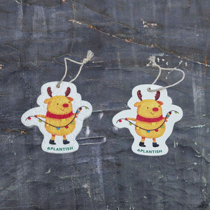 Top-view comparison of the Holiday pop-up sponge in dry versus wet states. The right image shows the sponge in its dry form - flat and compact, while the left demonstrates its expanded, thicker state after hydration. Both clearly feature the festive Holiday Moose design wearing a red scarf and holding string lights. Shot against a dark grey textured background, this eco-friendly kitchen cleaner showcases impressive size expansion when wet
