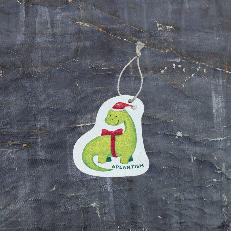 Top view of an eco-friendly cellulose pop-up kitchen sponge with hanging cord, featuring an adorable holiday dinosaur wearing a red Santa hat and red bow tie - lying flat when dry and expanding when wet for festive yet functional cleaning.