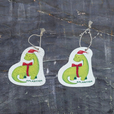 Top-view comparison of the holiday
dinosaur pop-up sponge in dry versus wet states. The right side shows the compact, flat disc in its dry form, while the left demonstrates the expanded, thicker cleaning pad after water absorption. Both feature an adorable holiday dinosaur wearing a red Santa hat with matching bowtie, complete with practical hanging cord. Shot against a dark grey textured background.