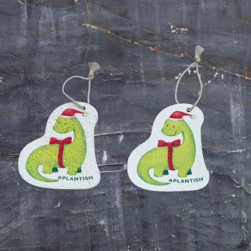 Top-view comparison of the holiday
dinosaur pop-up sponge in dry versus wet states. The right side shows the compact, flat disc in its dry form, while the left demonstrates the expanded, thicker cleaning pad after water absorption. Both feature an adorable holiday dinosaur wearing a red Santa hat with matching bowtie, complete with practical hanging cord. Shot against a dark grey textured background.