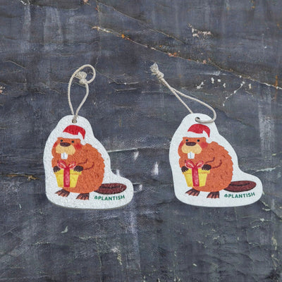 This top-view comparison showcases the holiday beaver pop-up sponge in both dry and hydrated states. On the right, the sponge appears as a flat, compact disc in its dry form, while the left displays its expanded, thicker cleaning pad state after water absorption. Both versions clearly showcase the adorable holiday beaver design complete with Santa hat and gift, along with a convenient hanging string. Photographed against a dark grey textured background.