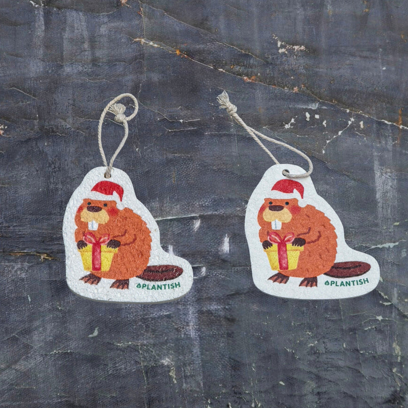 This top-view comparison showcases the holiday beaver pop-up sponge in both dry and hydrated states. On the right, the sponge appears as a flat, compact disc in its dry form, while the left displays its expanded, thicker cleaning pad state after water absorption. Both versions clearly showcase the adorable holiday beaver design complete with Santa hat and gift, along with a convenient hanging string. Photographed against a dark grey textured background.