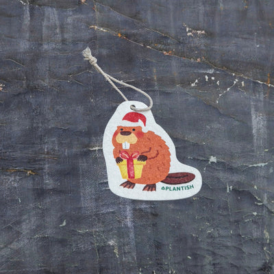 Top view of an eco-friendly cellulose pop-up kitchen sponge with hanging cord, featuring an adorable holiday beaver design wearing a red Santa hat and holding a red gift. This sustainable cleaning tool transforms from completely flat when dry to fully expanded when wet, combining festive cheer with practical functionality. Made from biodegradable plant-based cellulose, the seasonal sponge adds holiday spirit to your kitchen routine while being environmentally responsible.