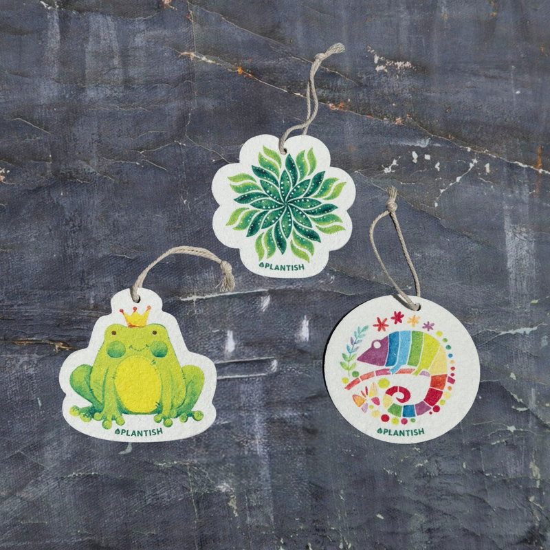 A top-view display of three eco-friendly pop-up sponges featuring nature-inspired designs. This set includes: 1) Frog, 2) Chameleon, and 3) Green Leaf patterns. These circular kitchen sponges are presented on a dark grey textured background. Made from biodegradable cellulose with attached strings, they bring the beauty of nature into your daily cleaning routine while being gentle on the environment.