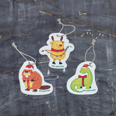 Top view of three eco-friendly pop-up sponges featuring festive holiday animal designs. This charming set includes: 1) Holiday Moose, 2) Holiday Beaver, and 3) Holiday Dinosaur patterns. Displayed on a dark grey textured background, these circular kitchen sponges are crafted from biodegradable cellulose with attached strings, bringing cheerful holiday spirit to your cleaning routine while being kind to the environment