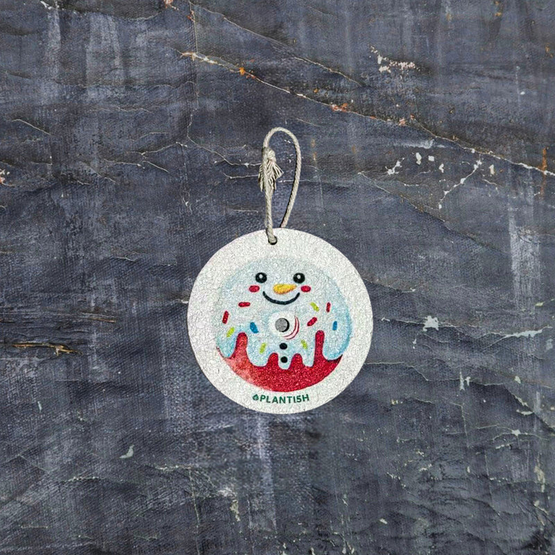 Top-down view of an eco-friendly cellulose pop-up kitchen sponge with hanging string, flat when dry and expands when wet, featuring a cute snowman donut design.