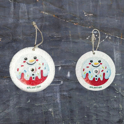 Top-view comparison of the Snowman Donut themed pop-up sponge in dry vs. wet states. On the left (dry): a flat beige disc displaying the snowman donut design with attached hanging string. On the right (wet): the expanded, thicker sponge maintains its circular shape with clearly visible pattern. Set against a dark grey textured background to highlight the product, this eco-friendly kitchen cleaning tool demonstrates significant volume increase when hydrated.