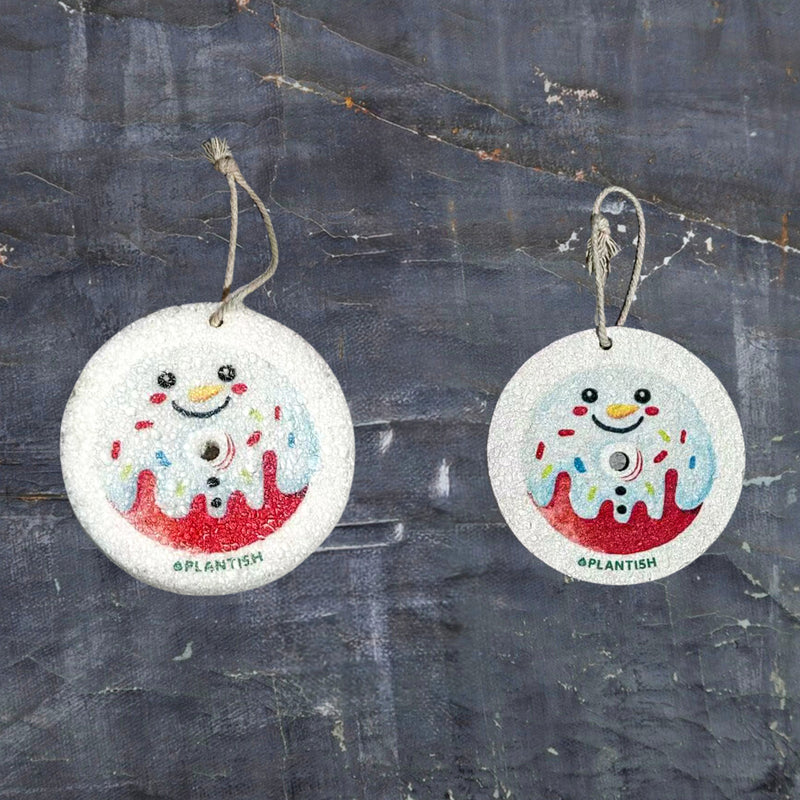 Top-view comparison of the Snowman Donut themed pop-up sponge in dry vs. wet states. On the left (dry): a flat beige disc displaying the snowman donut design with attached hanging string. On the right (wet): the expanded, thicker sponge maintains its circular shape with clearly visible pattern. Set against a dark grey textured background to highlight the product, this eco-friendly kitchen cleaning tool demonstrates significant volume increase when hydrated.