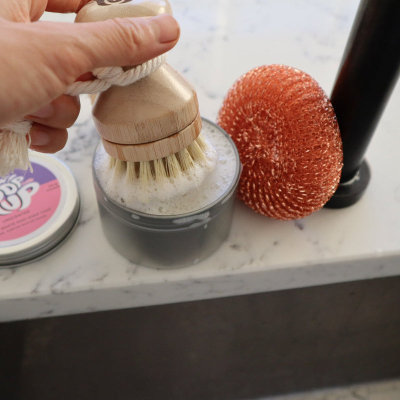 Hand holding a wooden scrubbing brush over a container of soap with a sponge and bottle in the background on a marble surface.