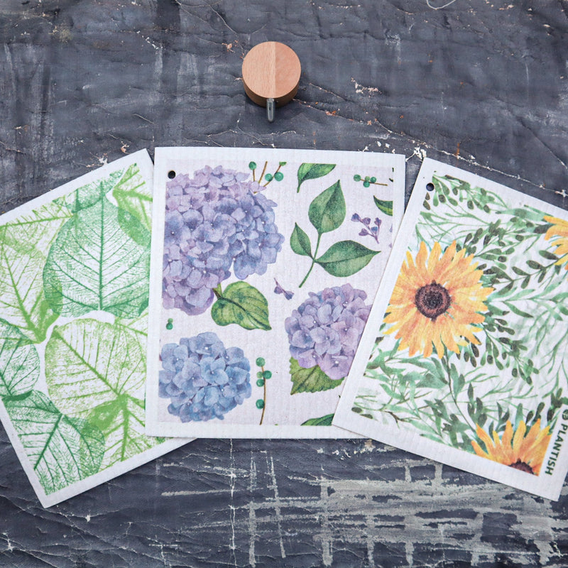 Three Sponge Cloths with floral and leaf patterns on a textured dark surface.