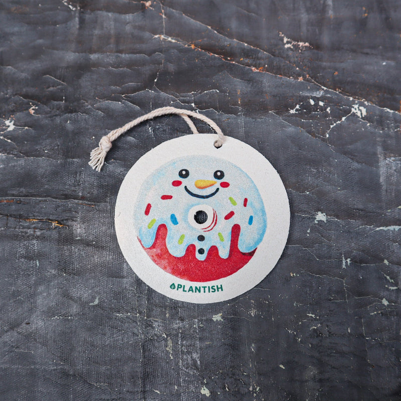 Top-down view of an eco-friendly cellulose pop-up kitchen sponge with hanging string, flat when dry and expands when wet, featuring a cute snowman donut design.