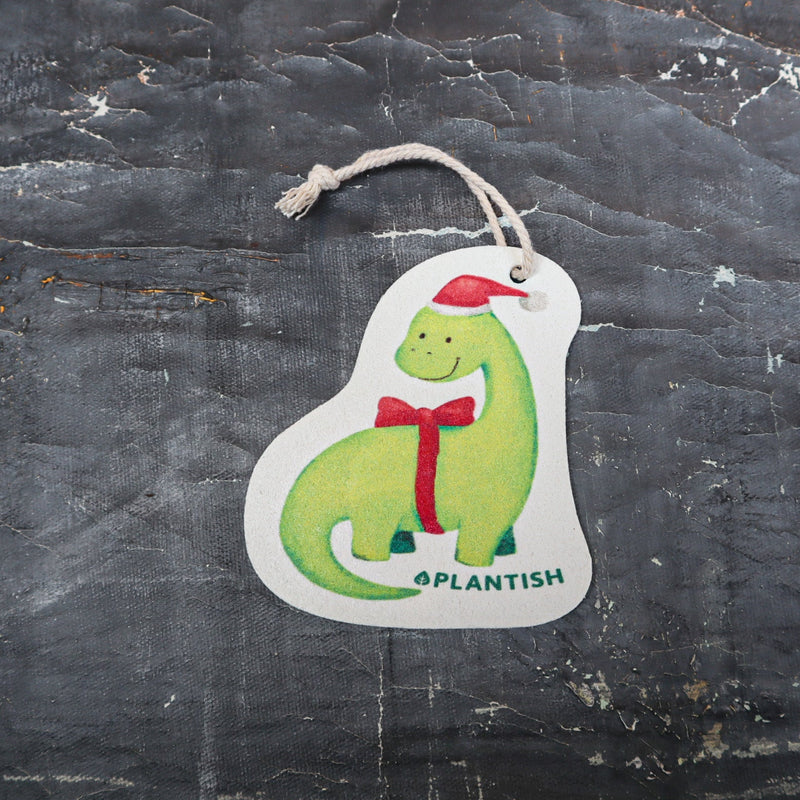 Top view of an eco-friendly cellulose pop-up kitchen sponge with hanging cord, featuring an adorable holiday dinosaur wearing a red Santa hat and red bow tie - lying flat when dry and expanding when wet for festive yet functional cleaning.