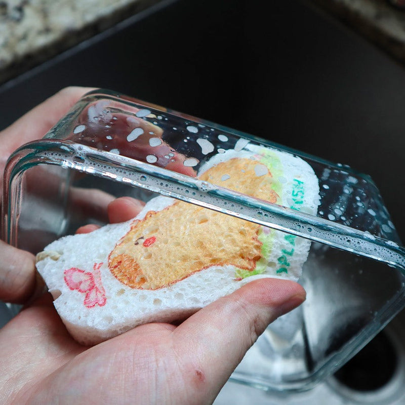 Eco-friendly pop-up sponge with capybara and butterfly design in action. Expanded cellulose kitchen sponge creating soap bubbles. Hand using biodegradable sponge inside clear glass rectangular bowl, demonstrating effective, sudsy cleaning. Playful, earth-friendly kitchen helper showing capybara with butterfly kiss on nose