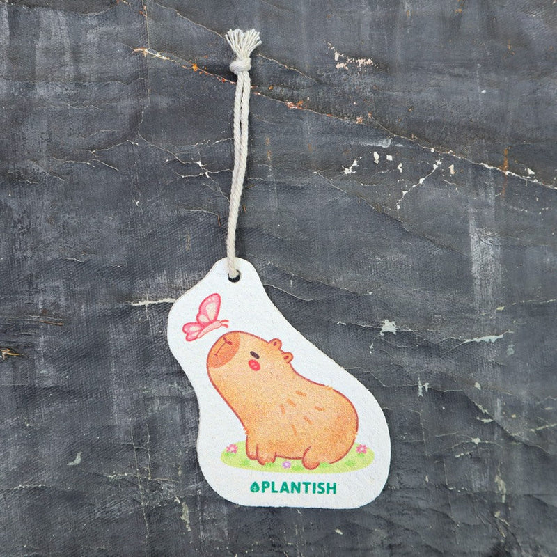 Top view of eco-friendly cellulose pop-up sponge for kitchen use. Flat, compact disc shape with thin string attached. Design features cute capybara with butterfly kissing its nose. Sponge expands when wet for effective cleaning. Natural beige background with visible cellulose texture. Capybara and butterfly illustrated in earthy tones.