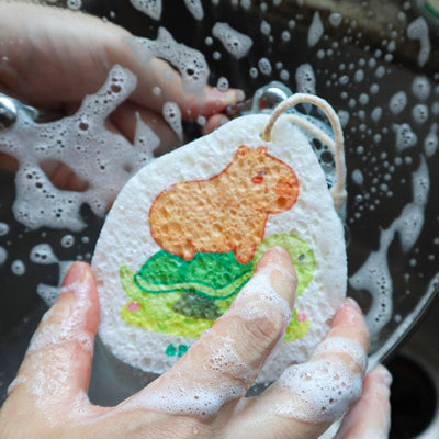 Eco-friendly pop-up sponge with capybara riding turtle design. Expanded cellulose kitchen sponge covered in soap bubbles. Hand using biodegradable sponge on dish, showing effective, sudsy cleaning. Playful, earth-friendly kitchen helper